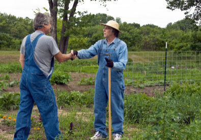 two men in bib overalls greet each other in a garden with a handshake