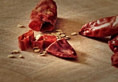 Sliced, dried, red serrano (Sport) chilli peppers on a chopping board with scattered chilli seeds. Knife marks are visible on the chopping board. Capsaicin, derived from chills, has medicinal properties, notably pain relief.